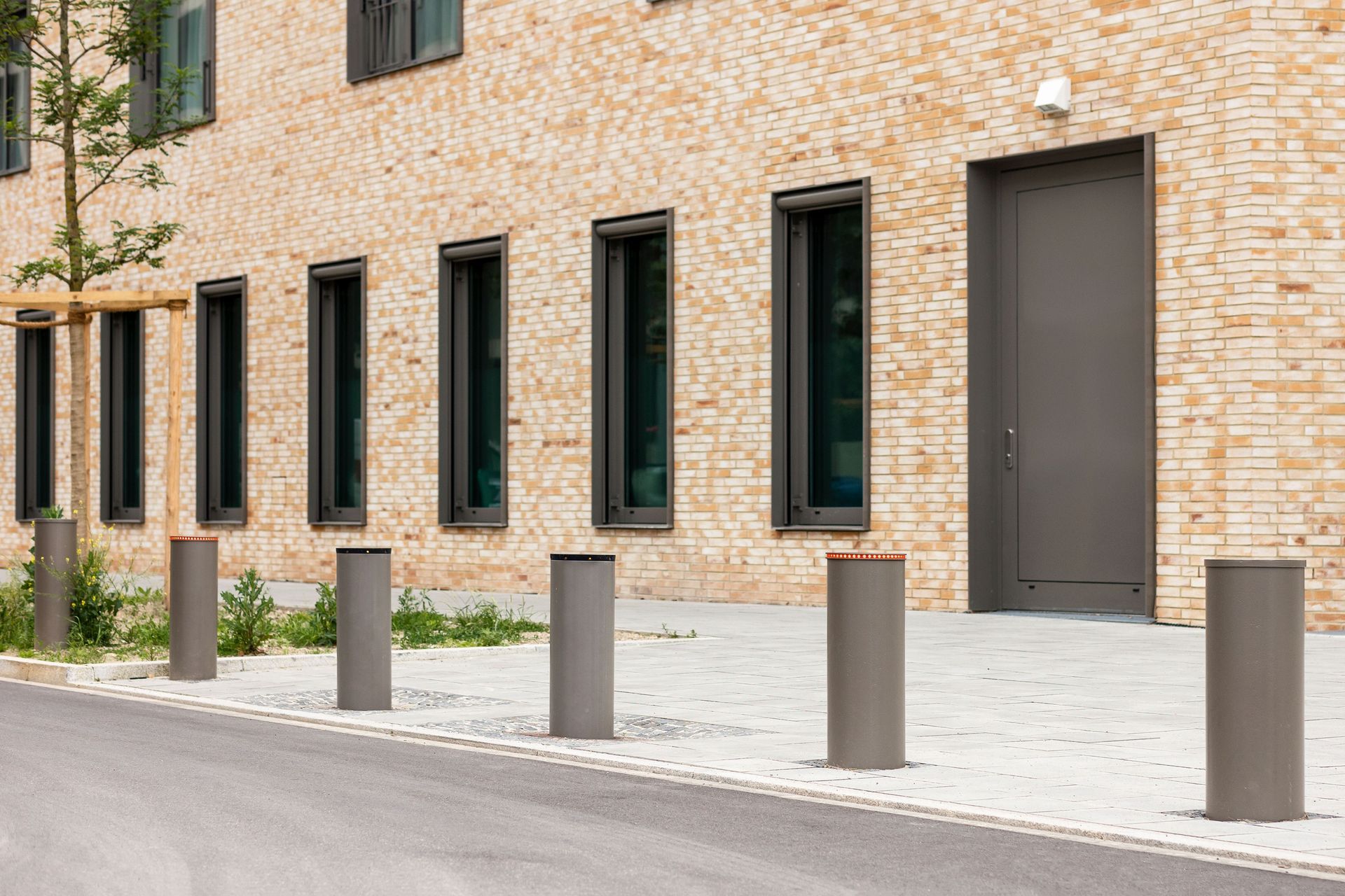 Brick apartment building with dark doors and windows, lined by gray bollards on a paved street