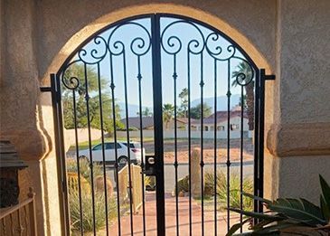Arched doorway with wrought-iron gate framing a sunny outdoor view of a parked car and homes beyond