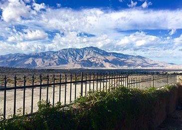 Mountain range under a blue sky with clouds, viewed past a black fence and green shrubs.