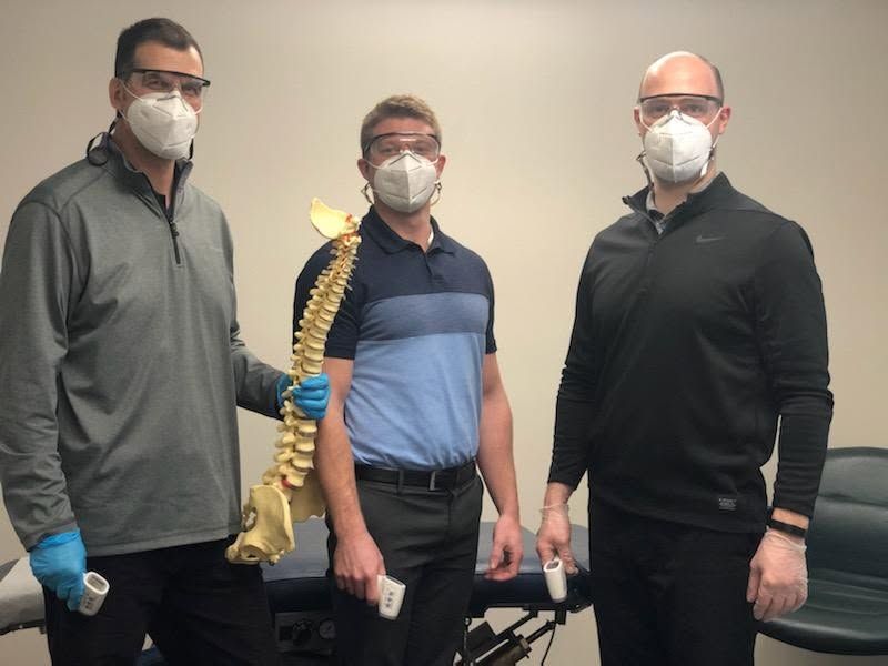 Three masked people stand beside a model spine in a clinic room.