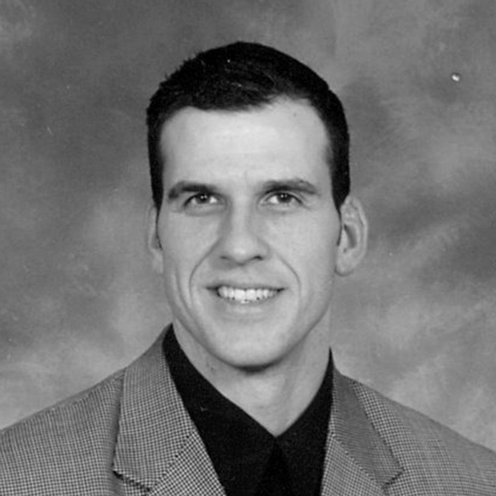 Studio portrait of a smiling man in a suit jacket and dark shirt against a gray backdrop