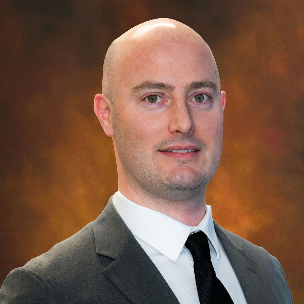 Studio headshot of a man in a dark suit and tie against a warm brown background