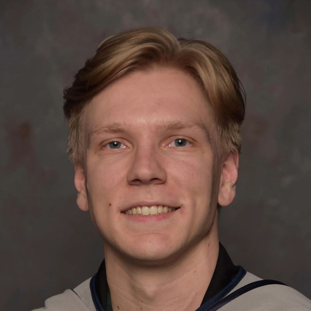 Portrait of a smiling person with short blond hair, wearing a light shirt against a dark gray background