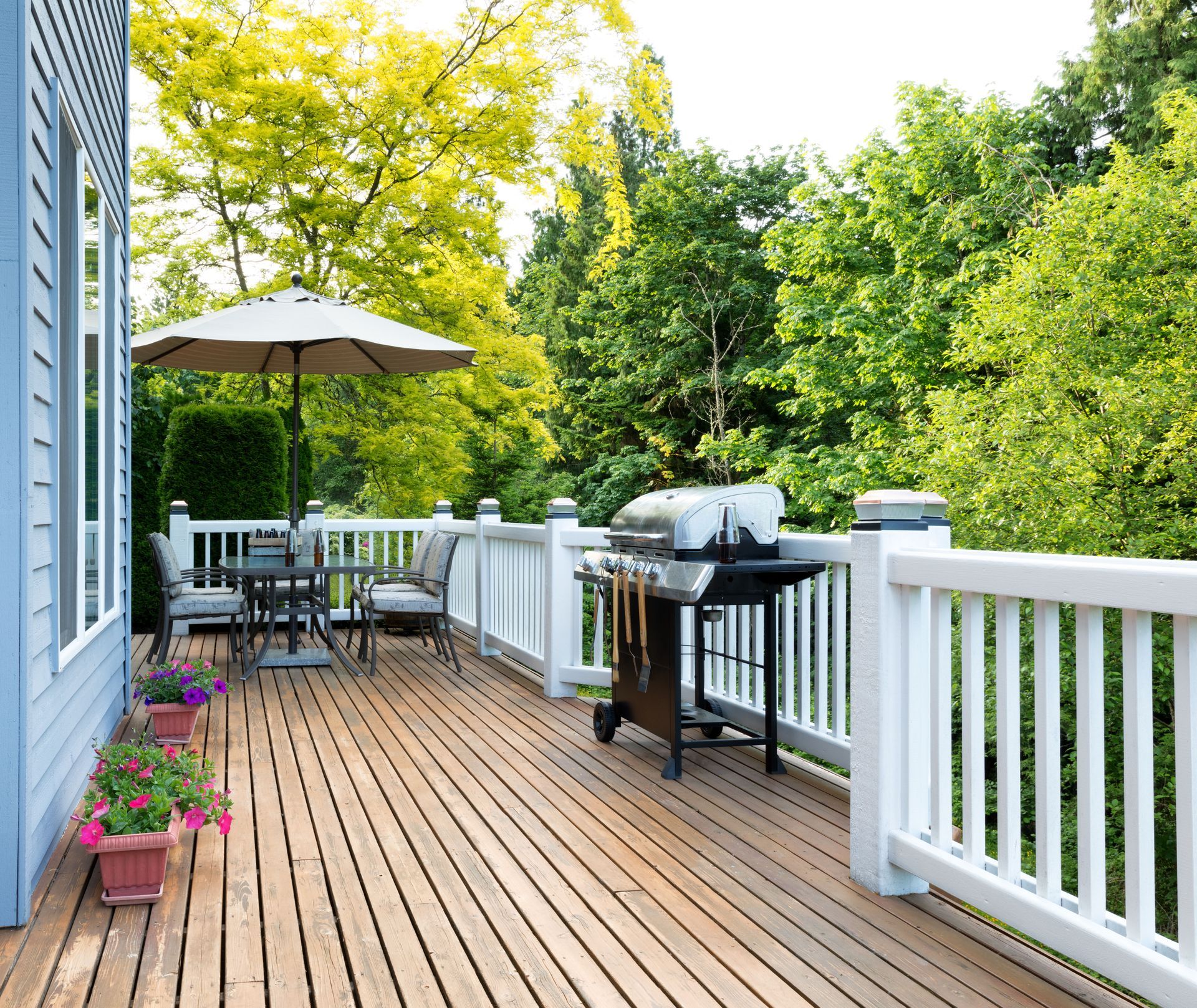 Wooden backyard deck with white railing, patio table, grill, umbrella, and potted flowers amid green trees