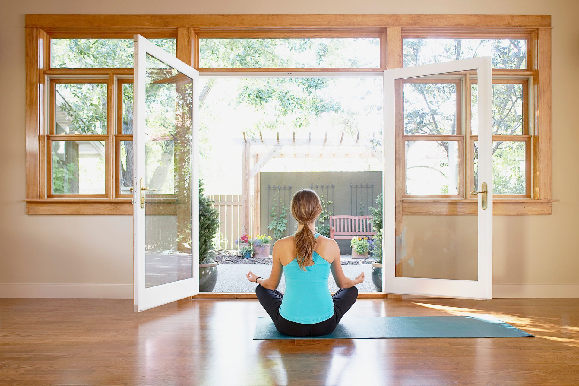 Person meditating on a mat in a bright room with open doors and windows looking out to a garden.