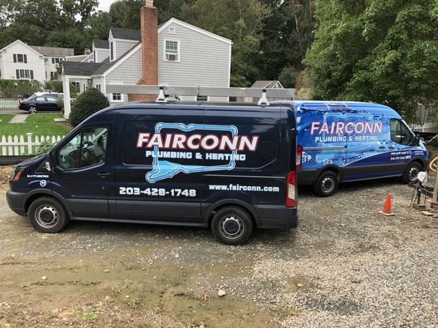 Two blue Fairconn Plumbing & Heating vans parked on a gravel lot near houses