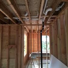 Partially framed interior of a house with exposed studs, ceiling pipes, and a window at the far end