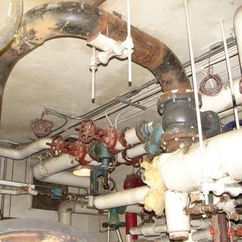 Industrial pipes and red valve wheels in a mechanical room with insulated ducts and overhead piping
