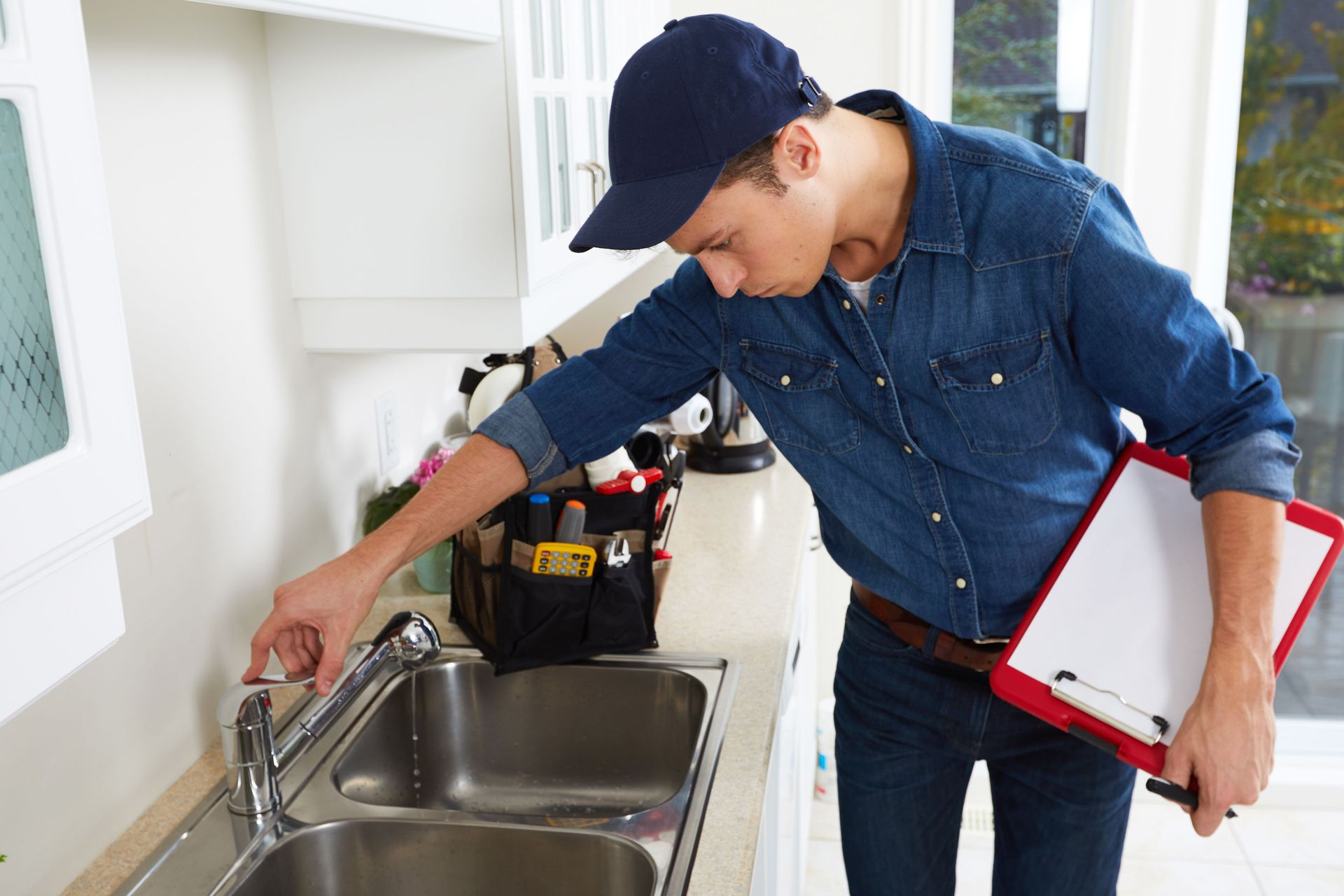 Plumber inspecting a kitchen sink faucet with a clipboard in a bright kitchen.
