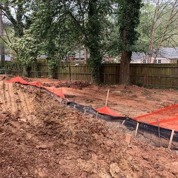 Construction site with red erosion control fencing around a muddy, cleared yard near trees and a wooden fence