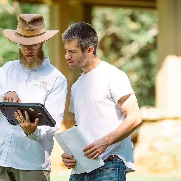 Two men reviewing a tablet outdoors; one holds papers, the other wears a hat and points at the screen.