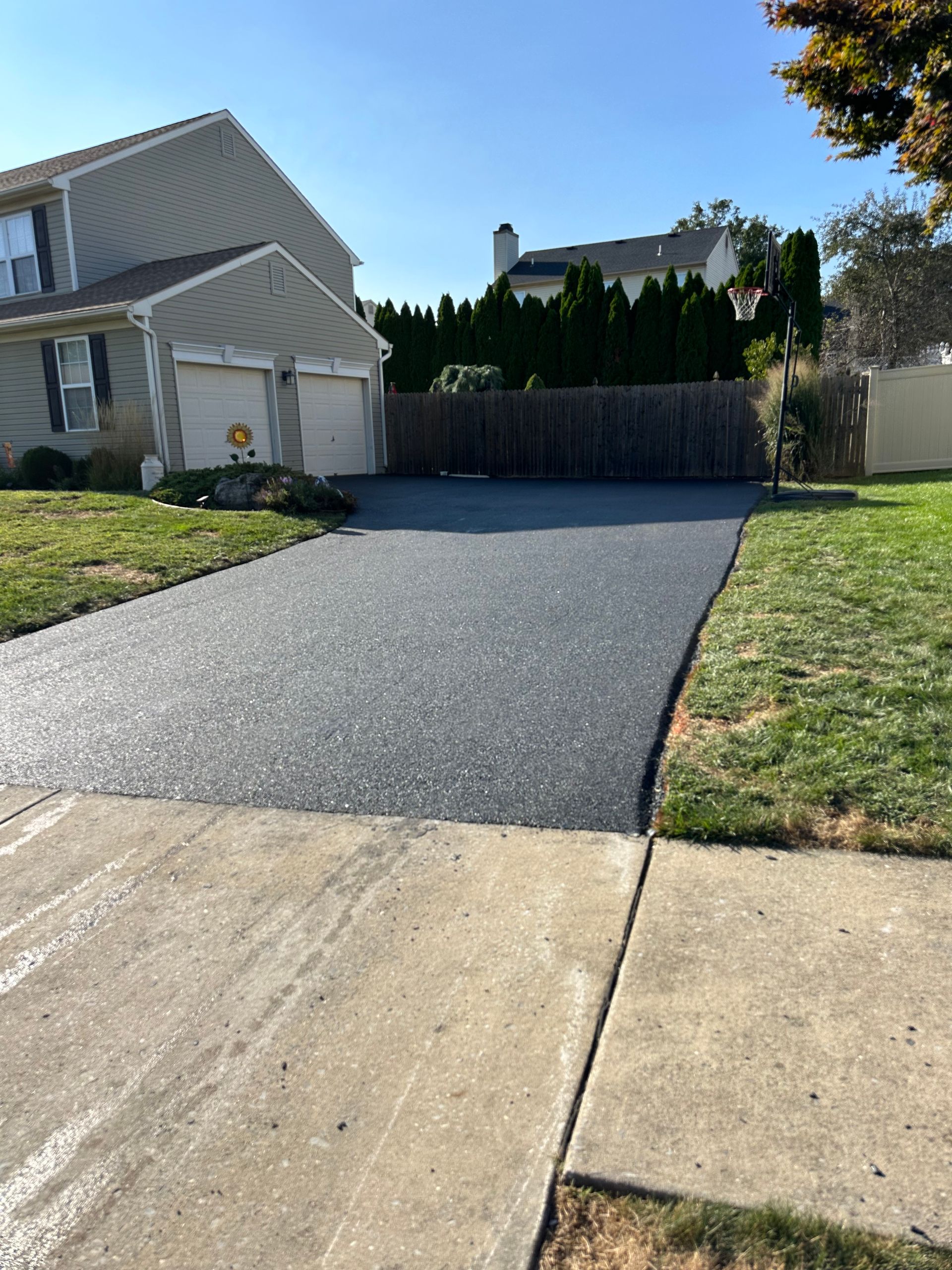 Fresh black asphalt driveway leading to a wooden gate beside a house and lawn under bright sun