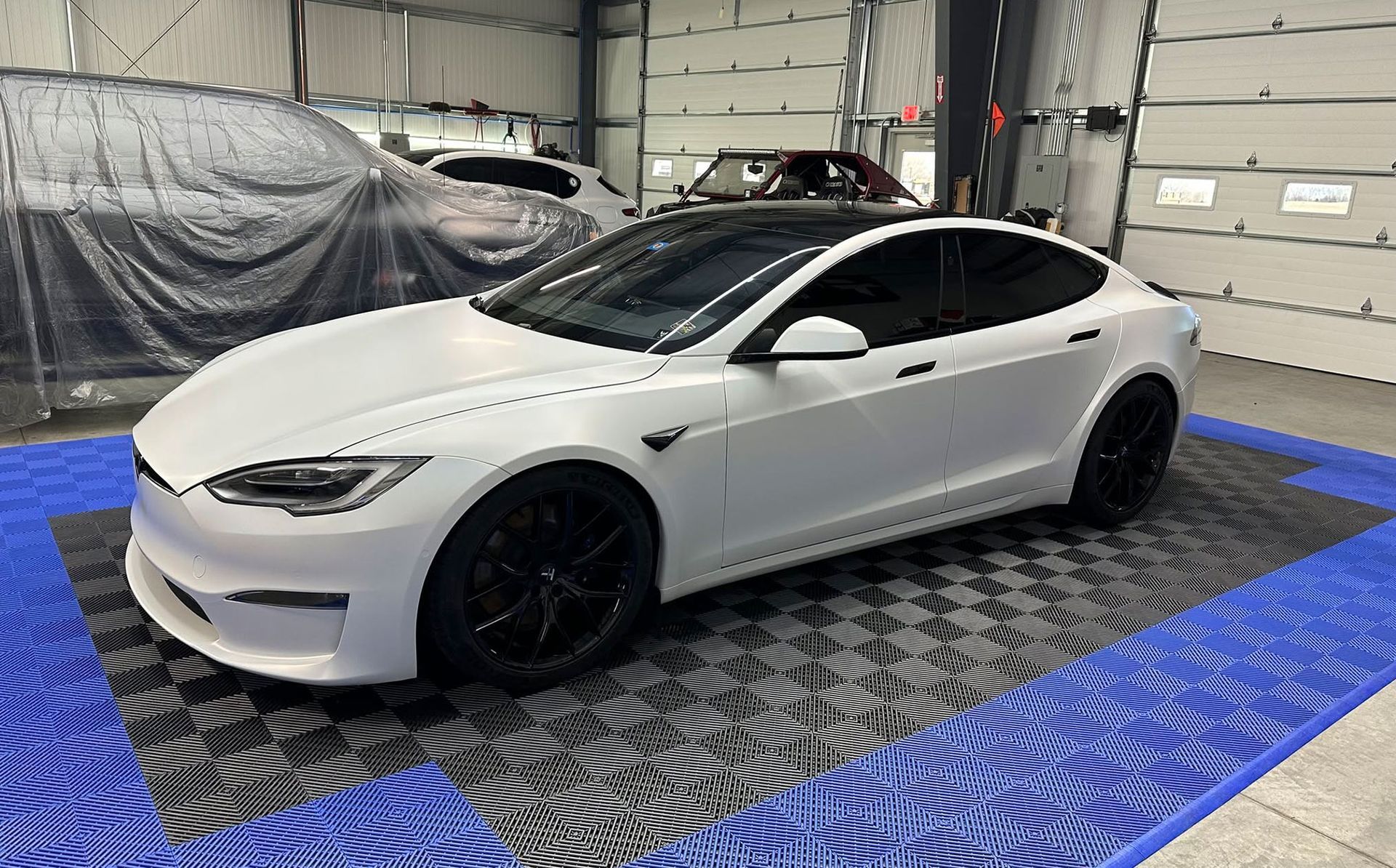 White Tesla sedan in a garage on a blue patterned floor mat, viewed from the front-left side