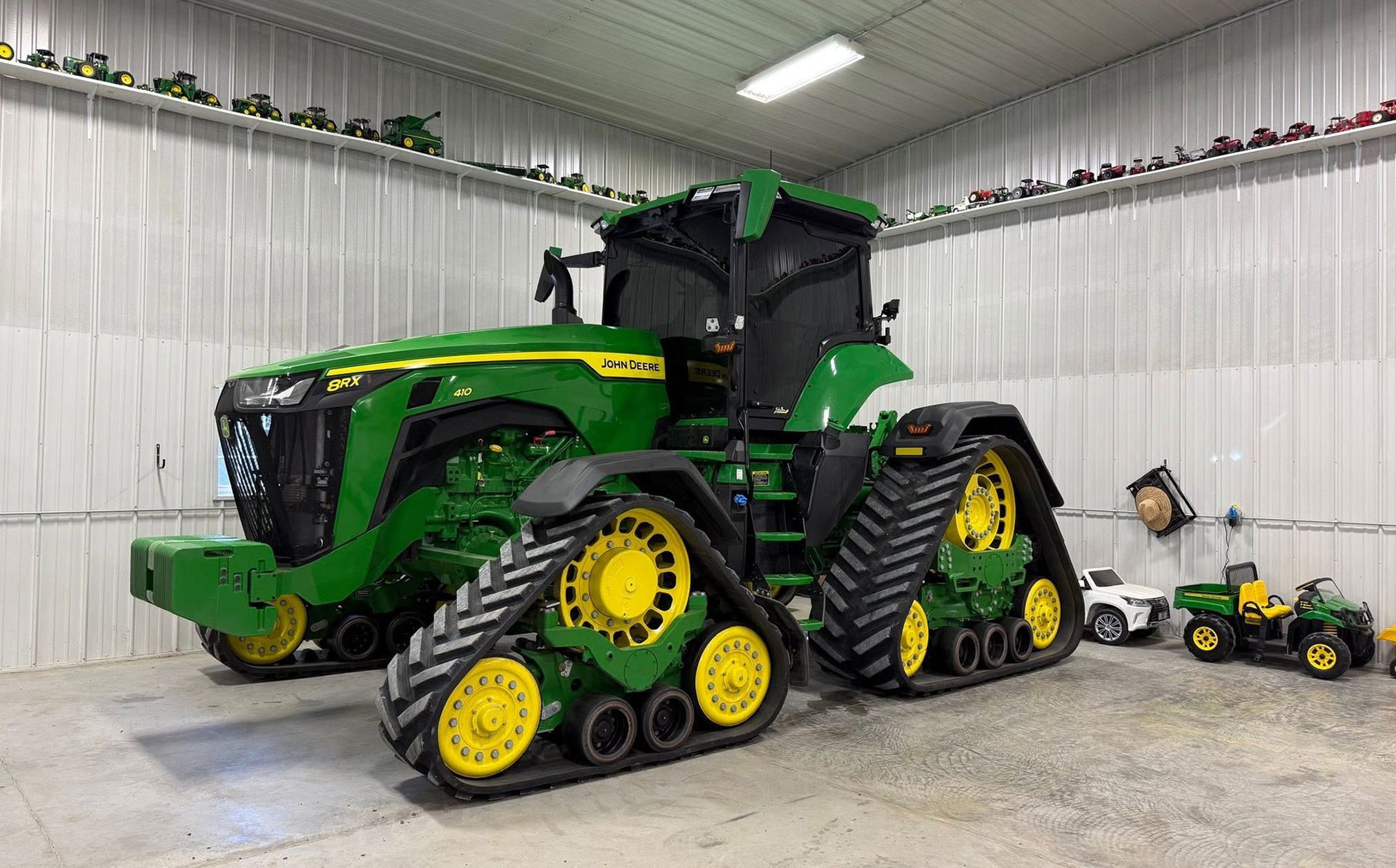 Green John Deere tractor with rubber tracks in a garage, beside two small toy tractors on the floor.