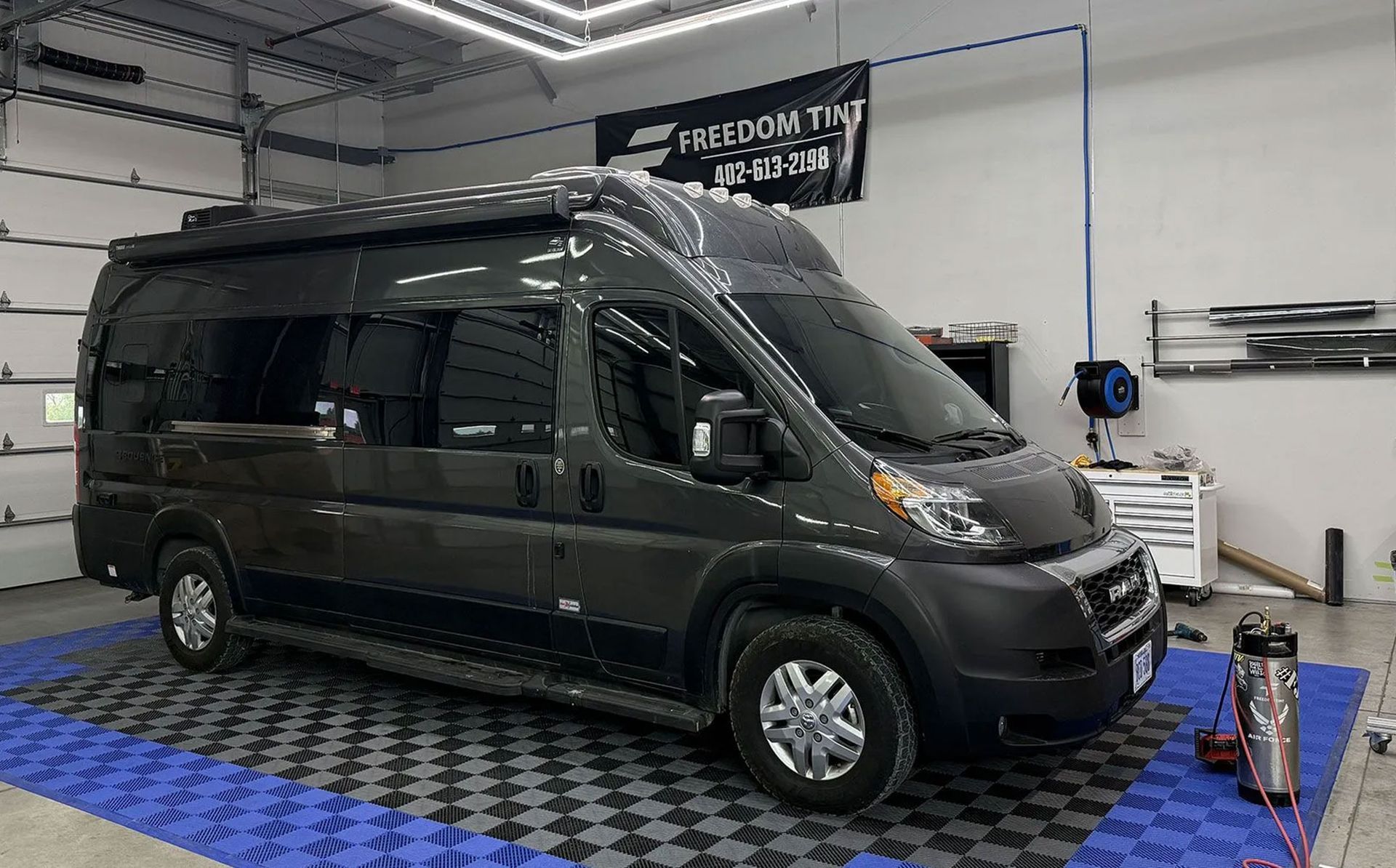 Black camper van in an indoor garage, parked on blue and black checkered flooring.