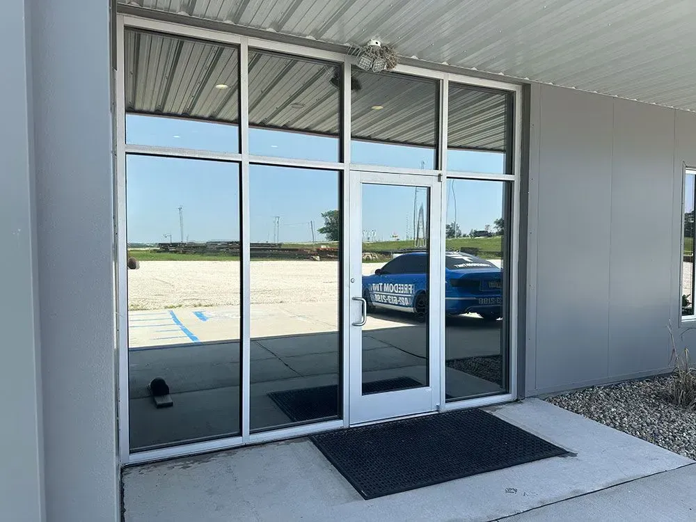 Glass storefront entrance with double doors, dark mats, and a blue car reflected outside
