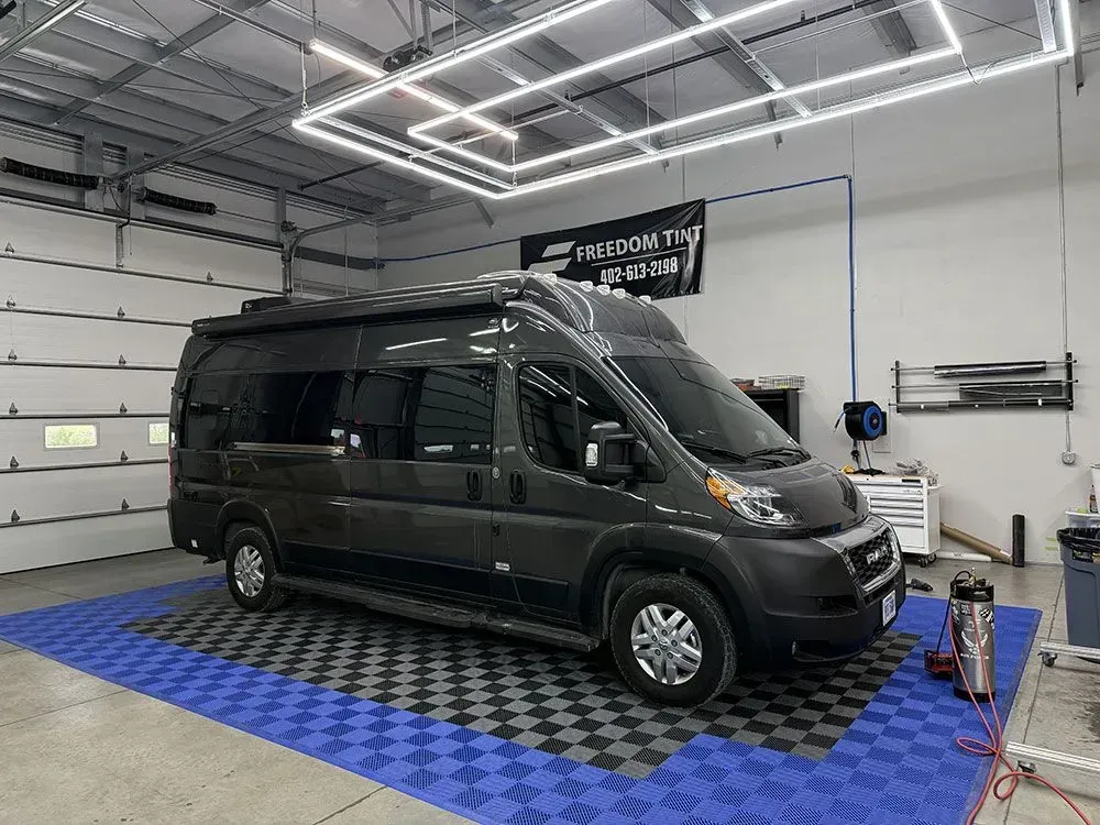 Black camper van in a garage on a blue checkered floor under bright overhead lights.