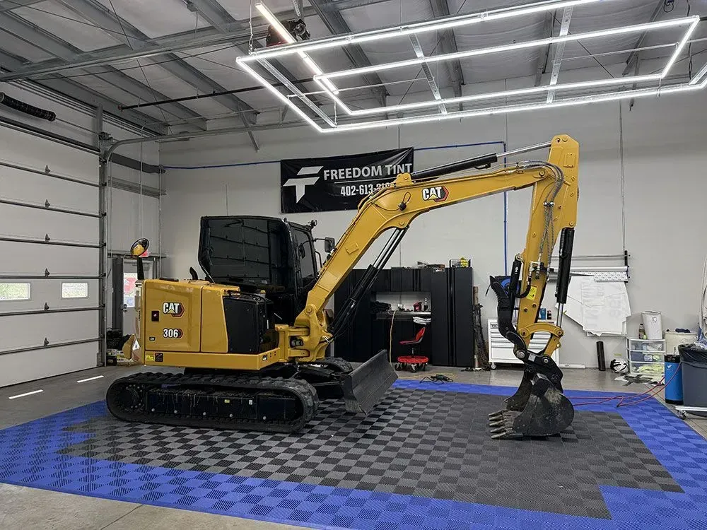 Yellow excavator in a workshop garage on checkered floor mats, holding a grapple attachment.