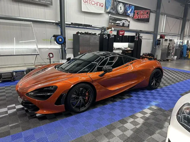 Orange sports car in a garage, shown from the front-left on a blue checkered floor.