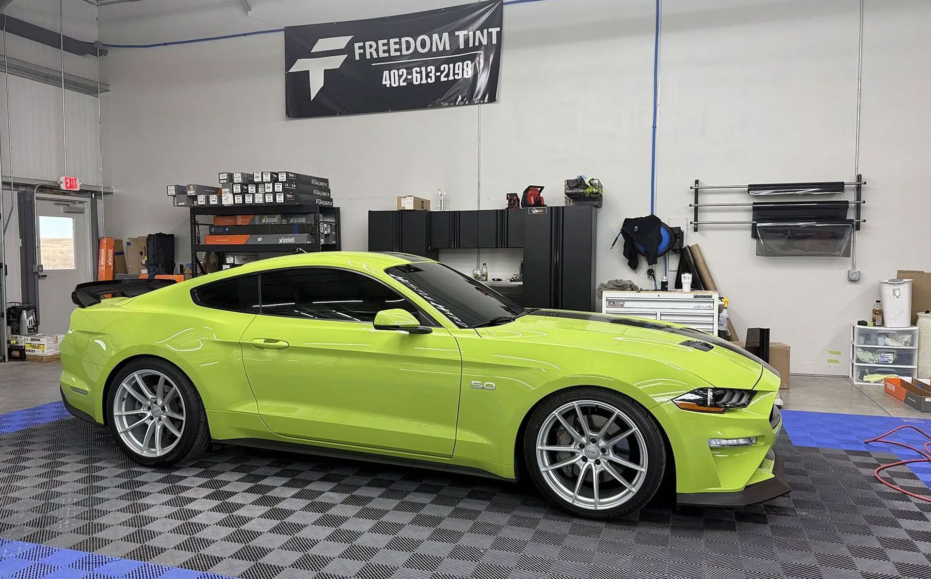 Lime green sports car in a garage, side view on a checkered floor with tools and shelves behind it.