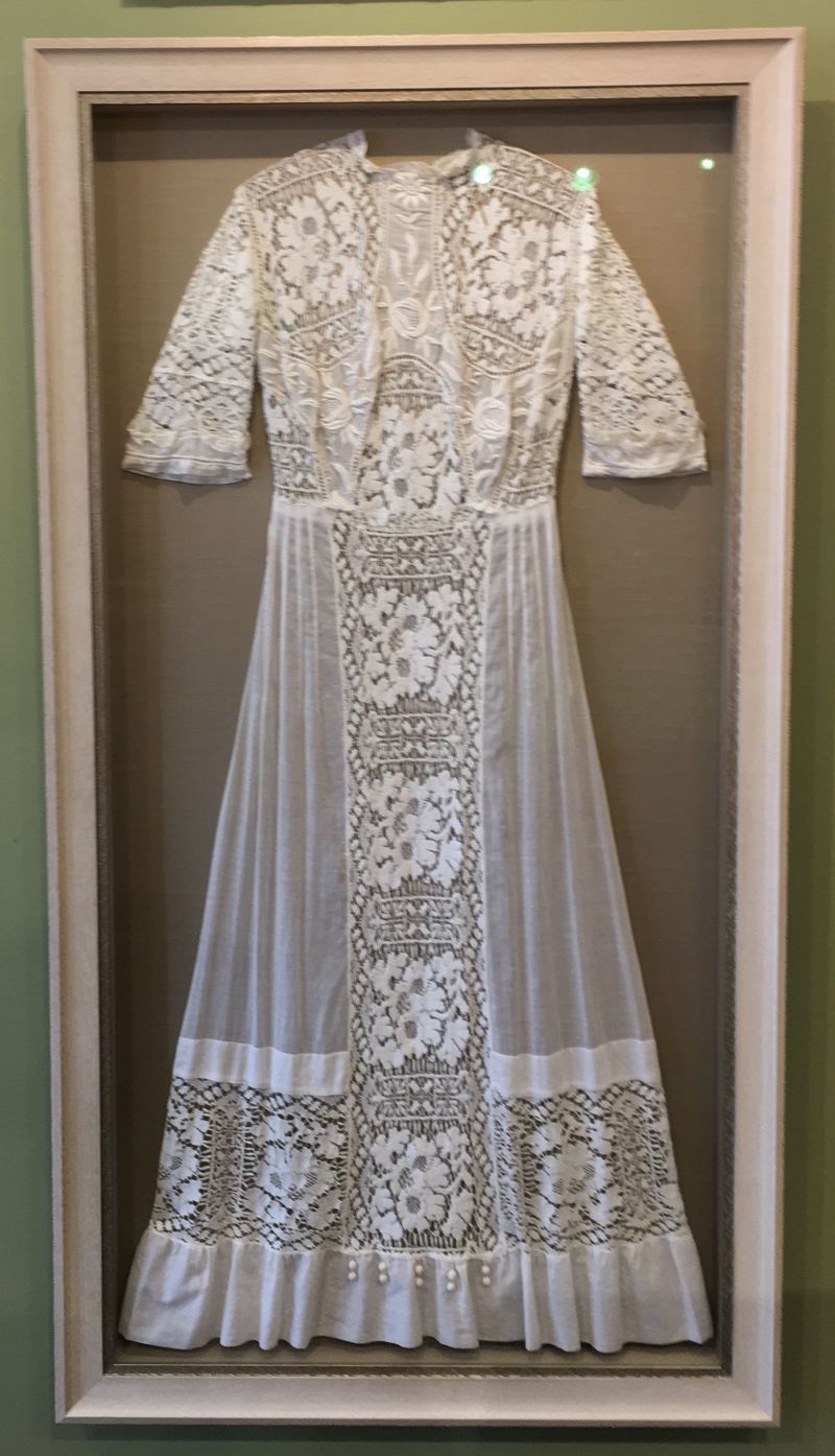 A white lace dress is in a frame on a wall.