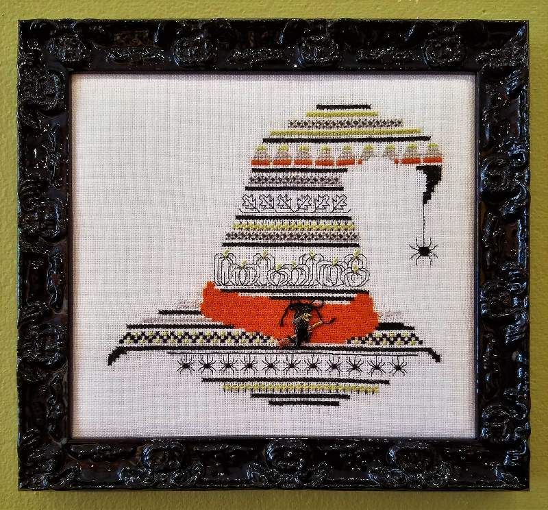 A framed cross stitch of a witch hat with a spider hanging from it