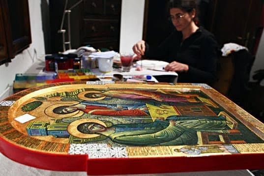 A person sits at a desk painting a large, ornate icon of three religious figures in a studio setting.