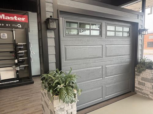 A grey residential garage door with window inserts, flanked by stone planters in a showroom setting.