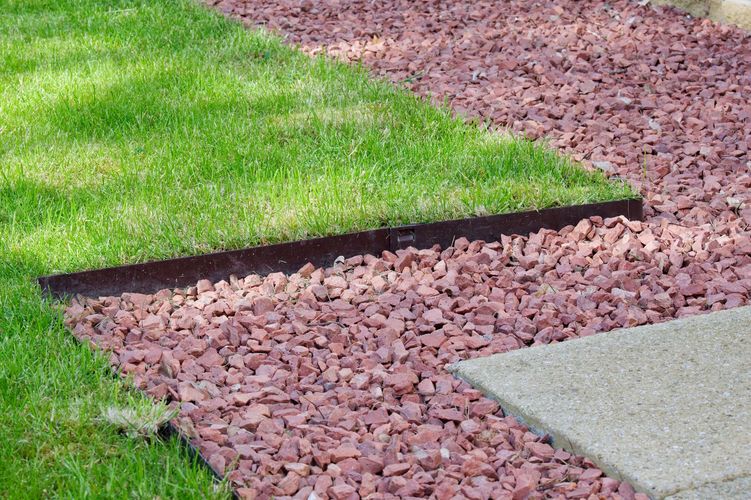 Curved garden edging separating green lawn, red gravel border, and a concrete walkway