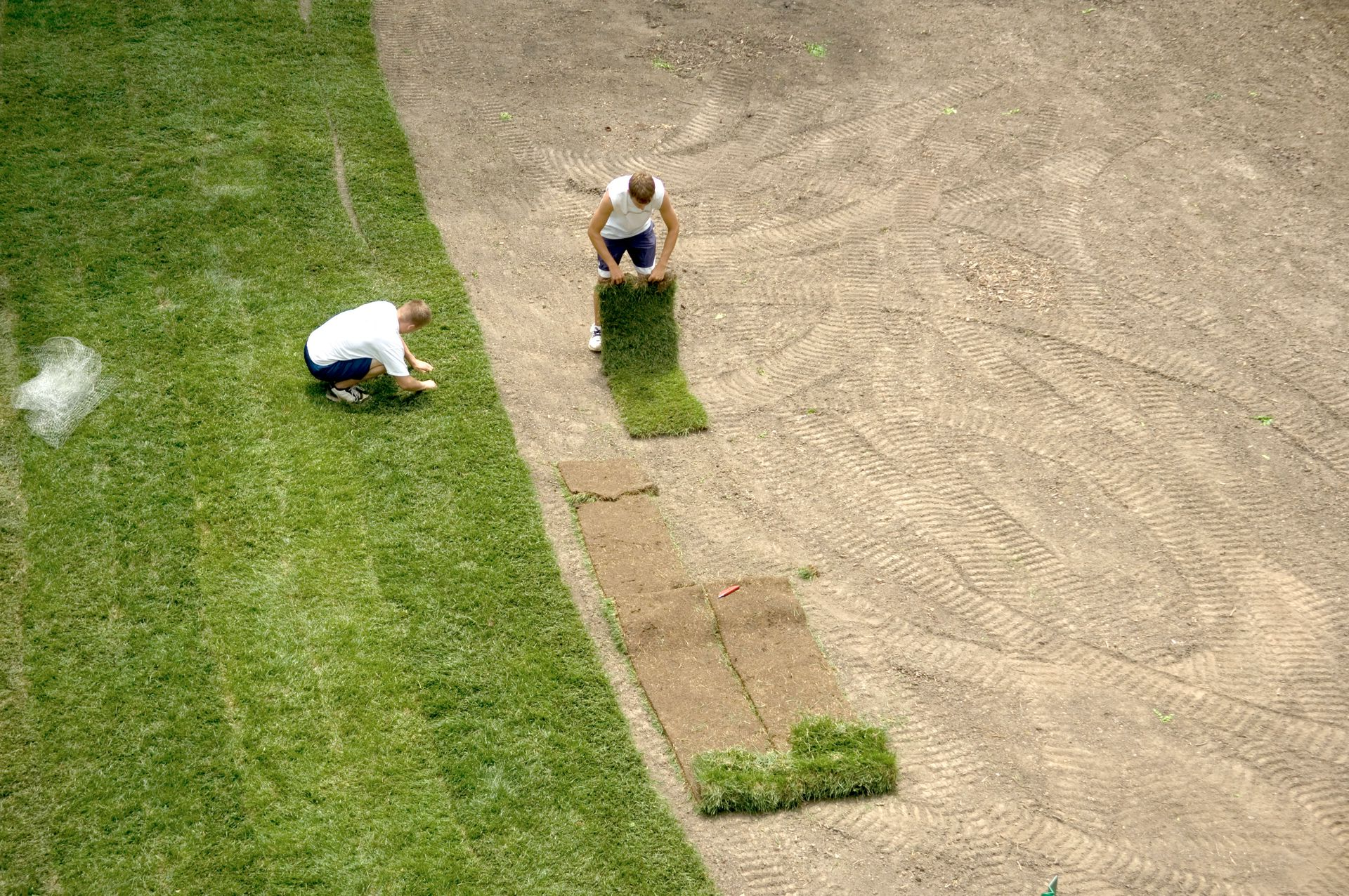 Two groundskeepers trimming grass beside a dirt path and cricket pitch