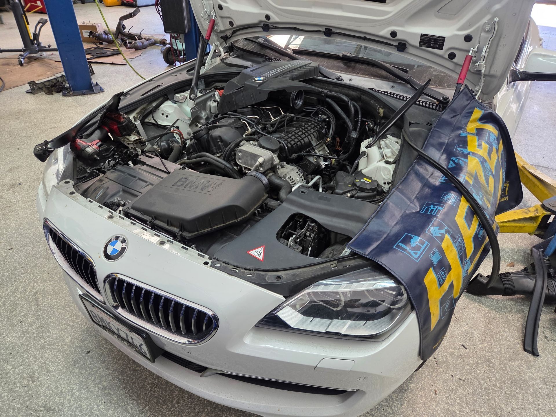 White BMW car with open hood in a repair shop, showing engine components.