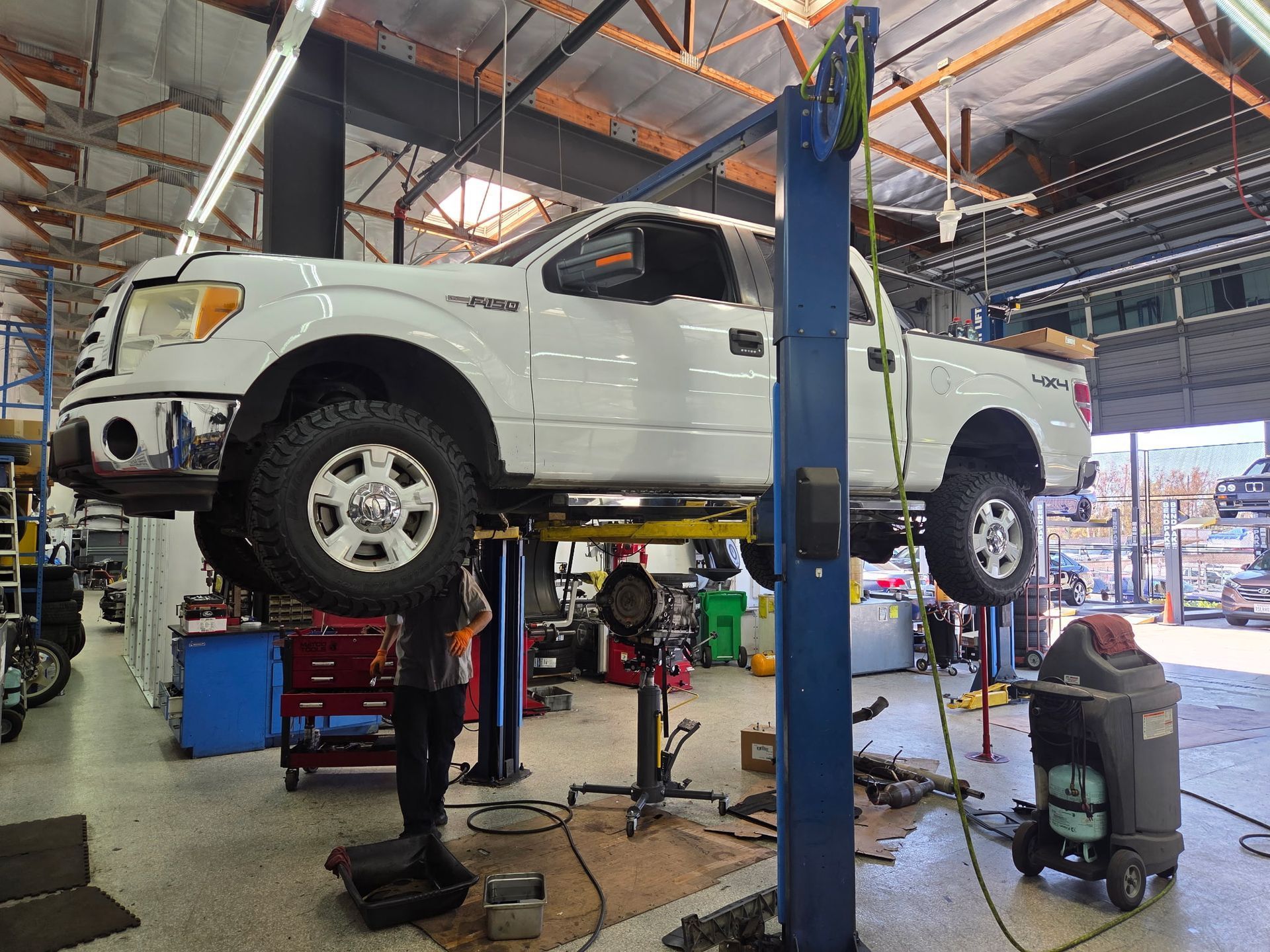 White pickup truck raised on a lift in a garage. Mechanic works below; various tools and equipment surround.