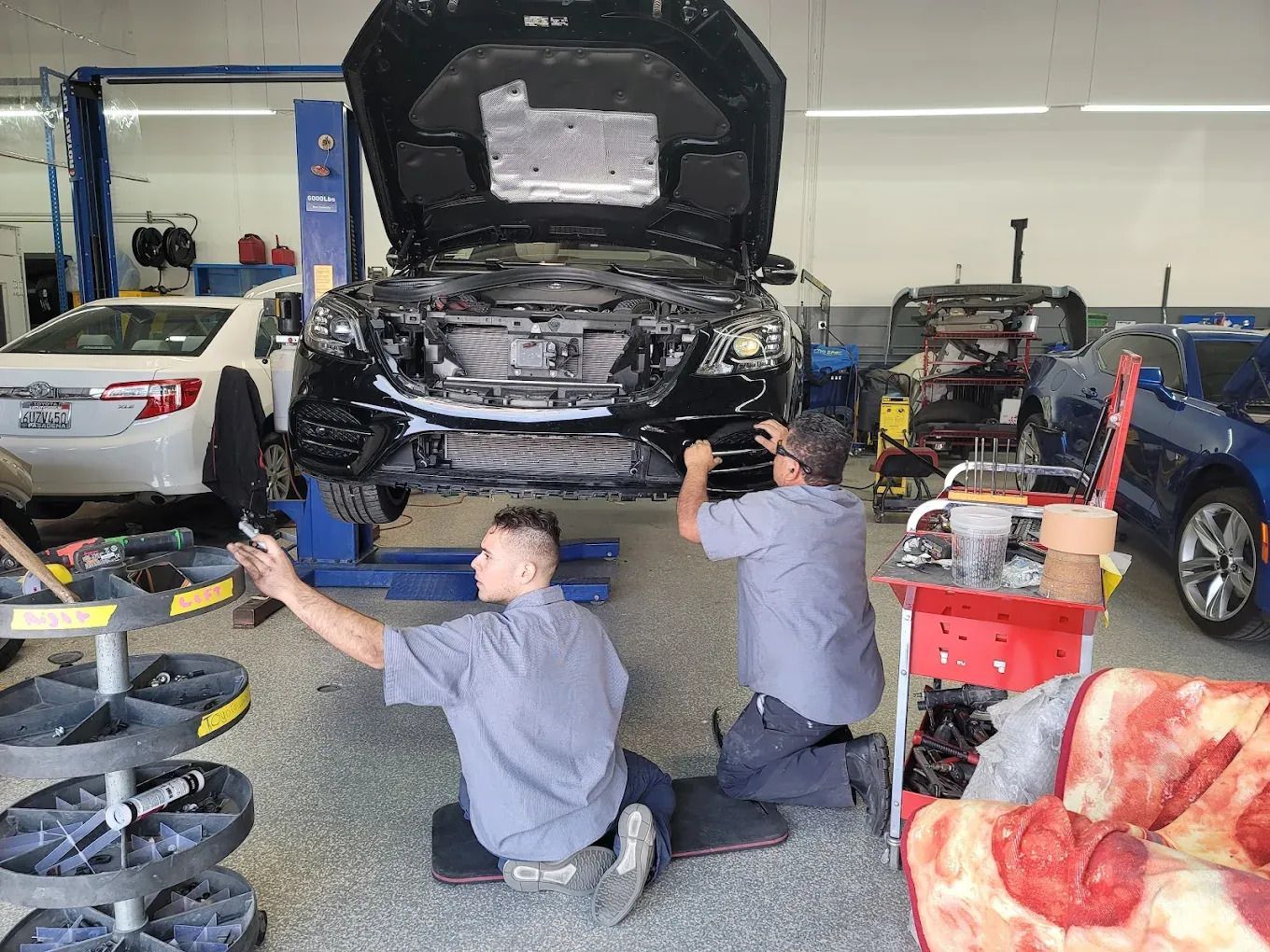 Two mechanics working on a black car with hood open in a garage, a white car and a blue car are in the background.