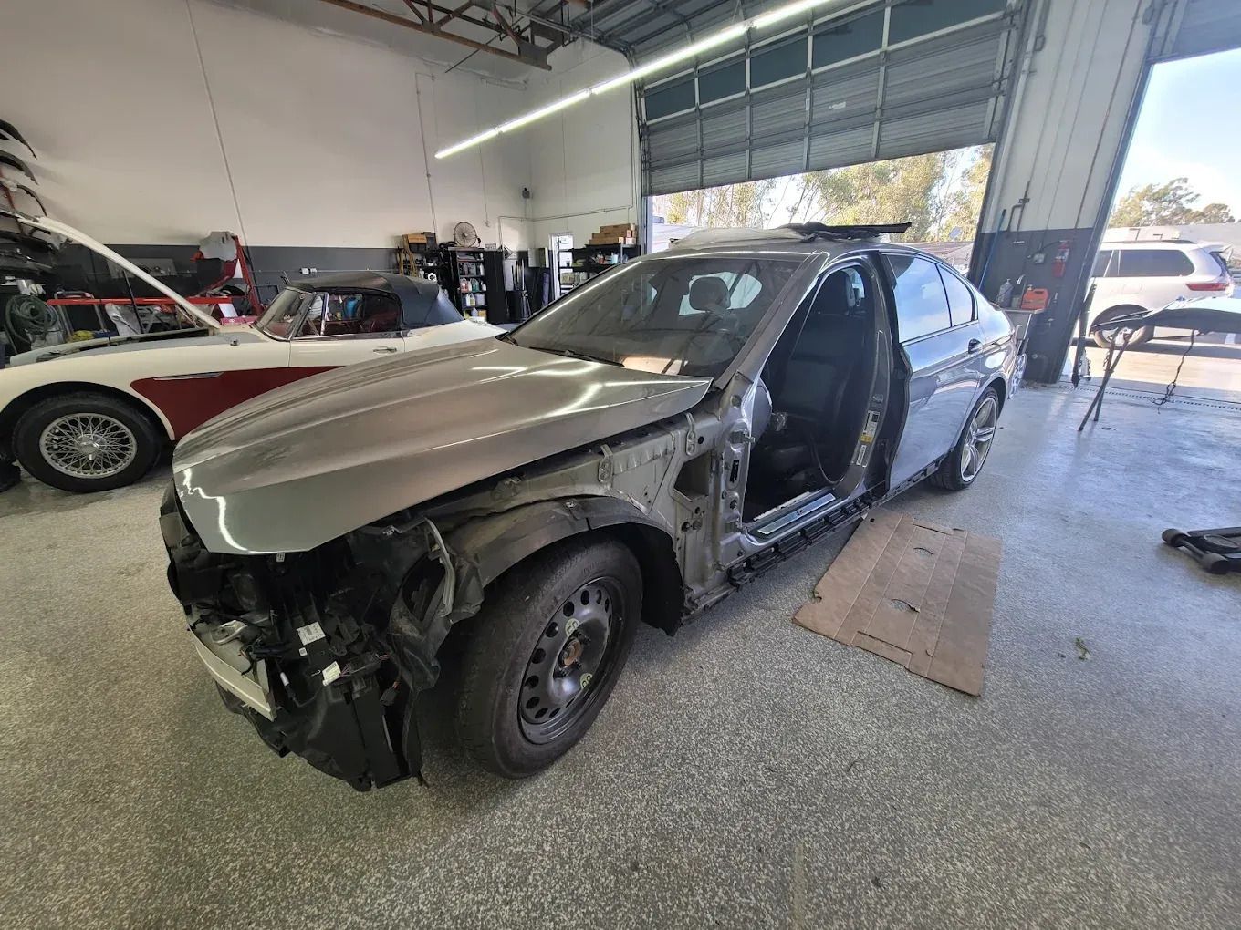 A gray car in a repair shop with its front and doors removed; a classic red car in the background.