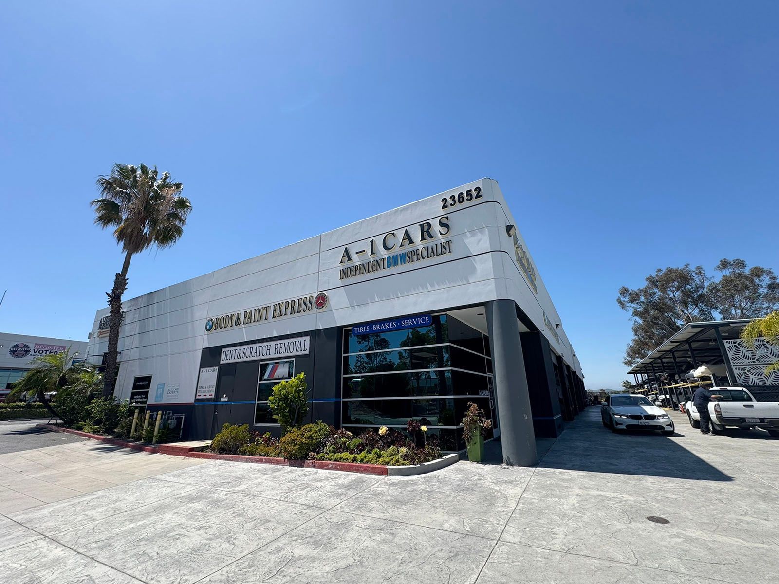 Modern white commercial building with large signage, blue sky, palm tree, and parked cars