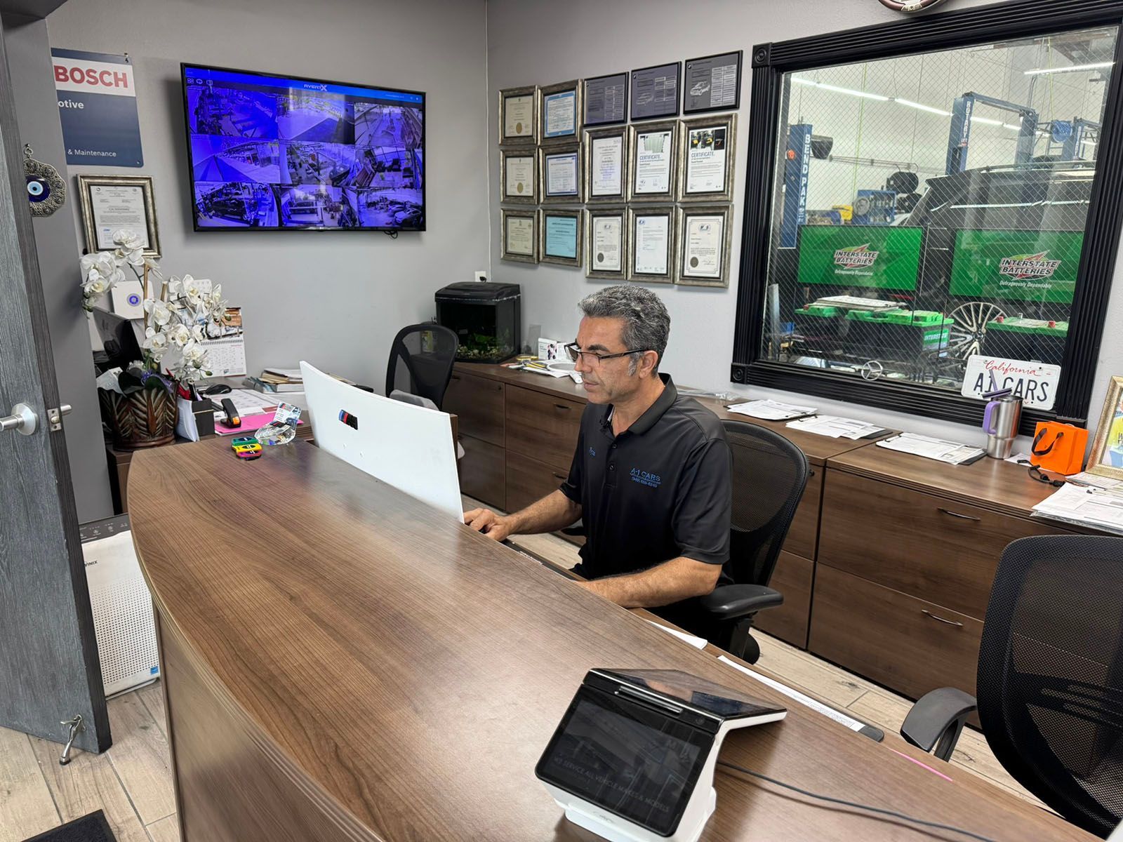 Office reception desk with man working on a laptop, framed certificates, and a wall-mounted TV in the background
