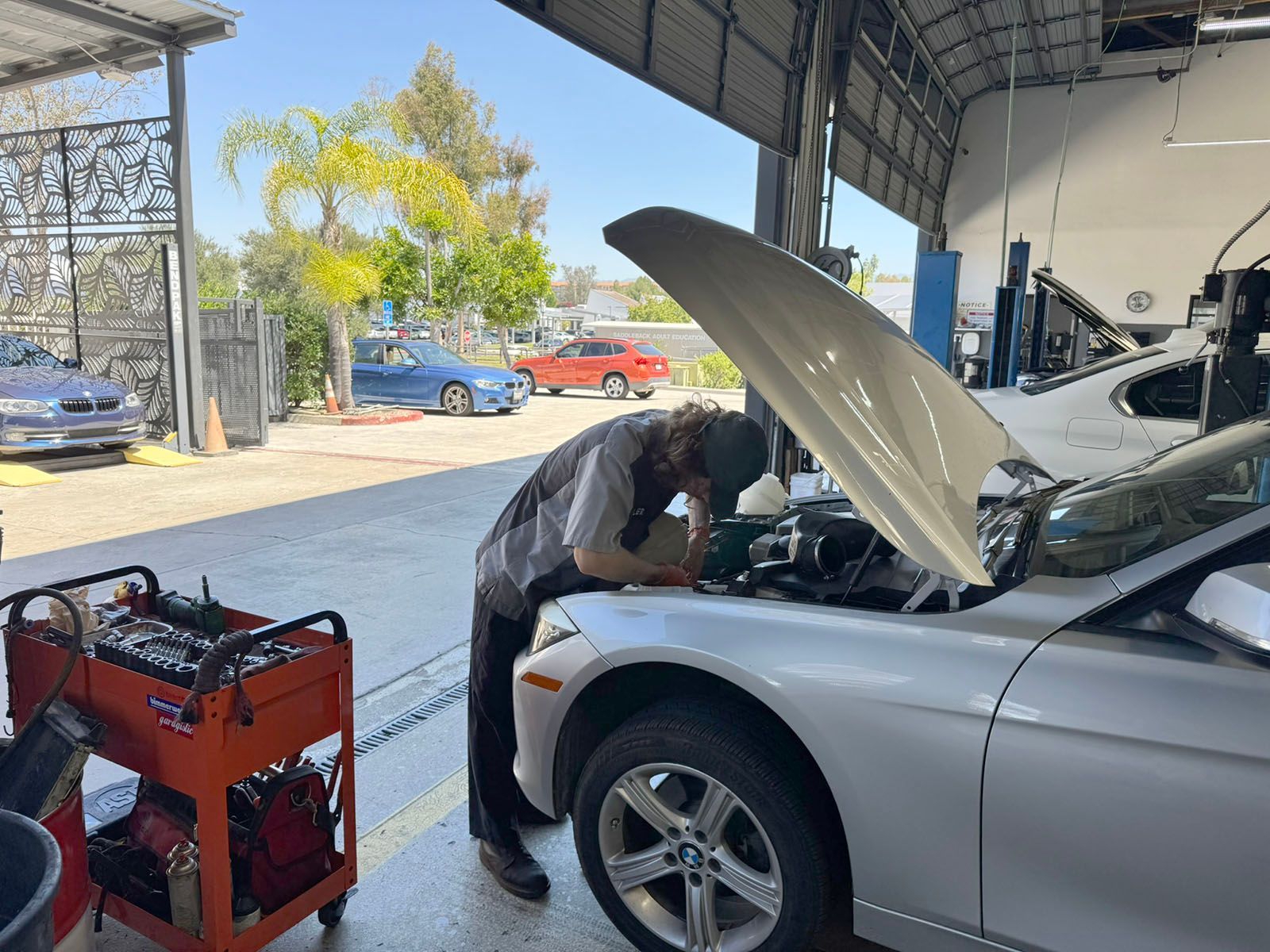 Mechanic working under open hood of silver car in a garage with red tool cart