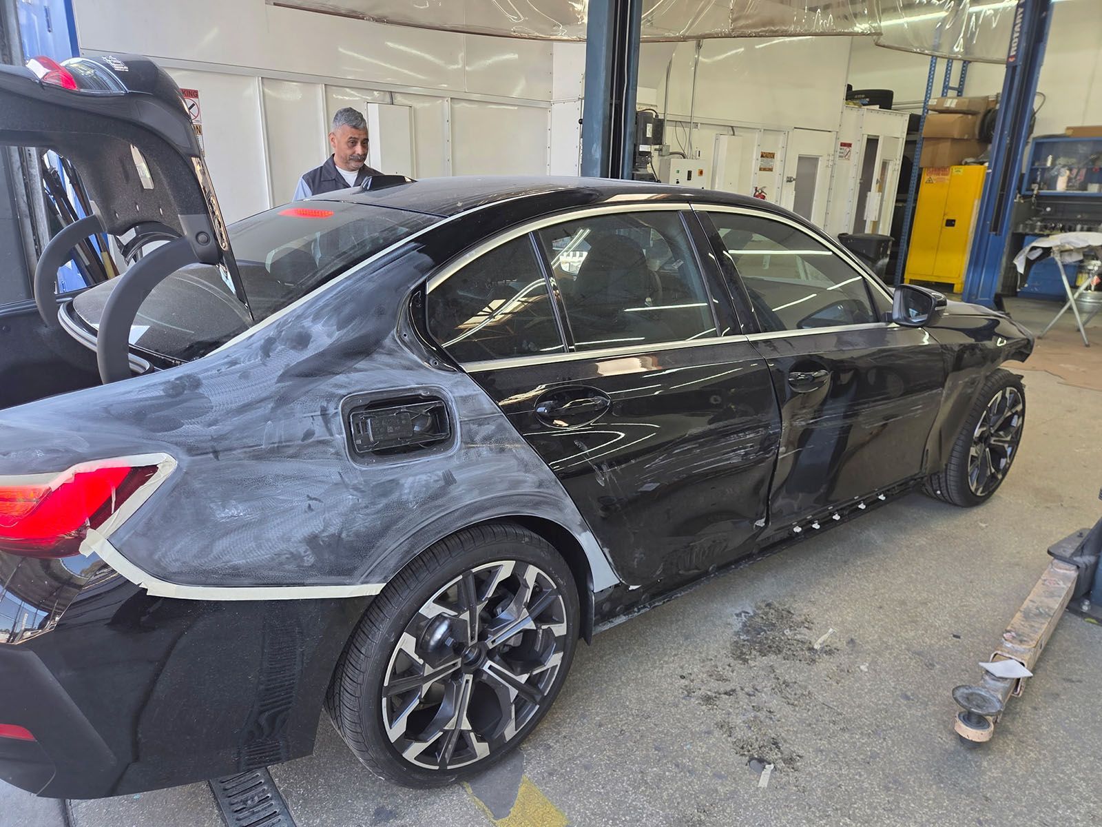 Black sedan with a damaged side panel in an auto repair shop, with a mechanic nearby