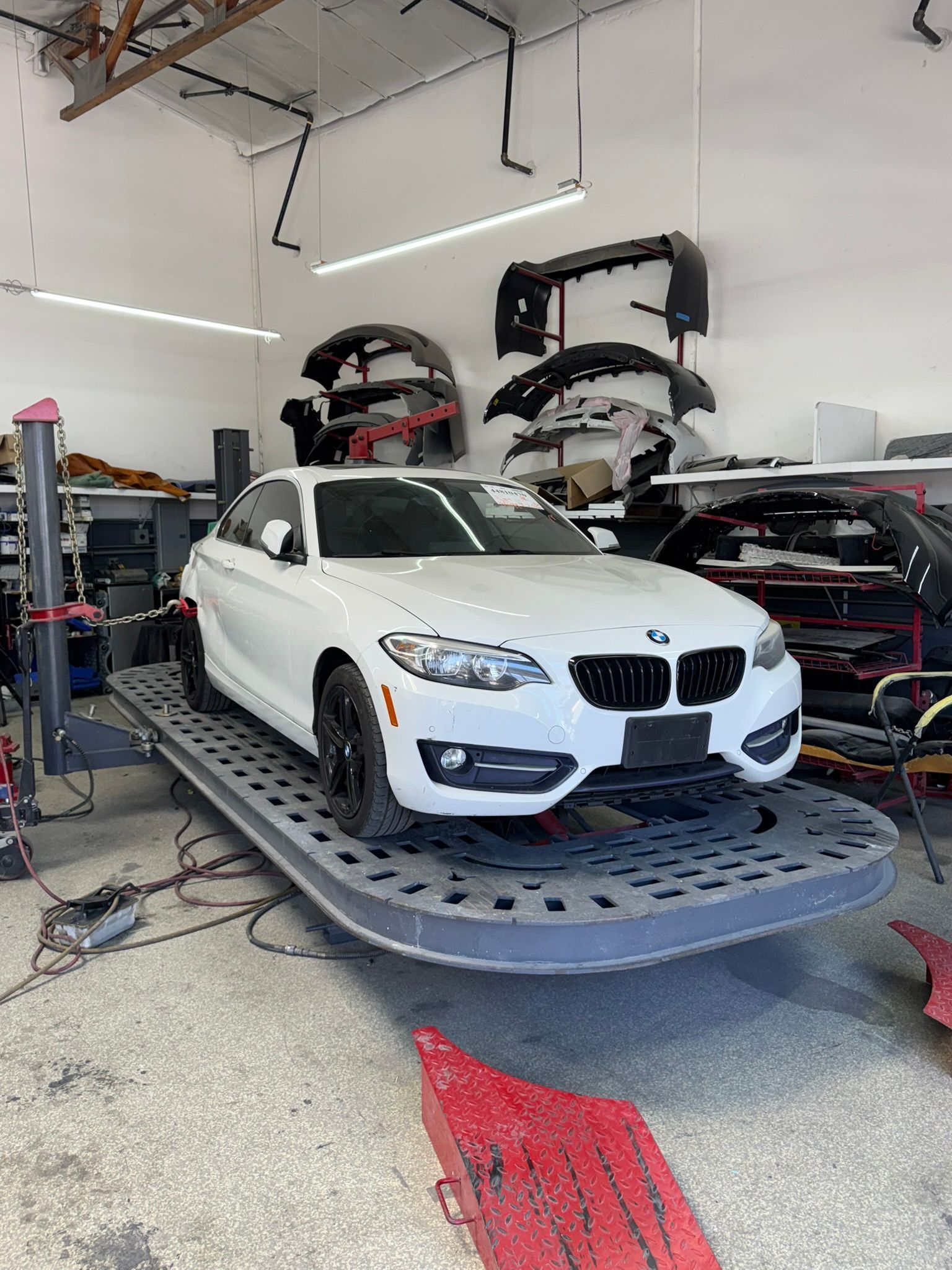 White BMW on a hydraulic lift in an auto shop, with other cars and shop equipment nearby.