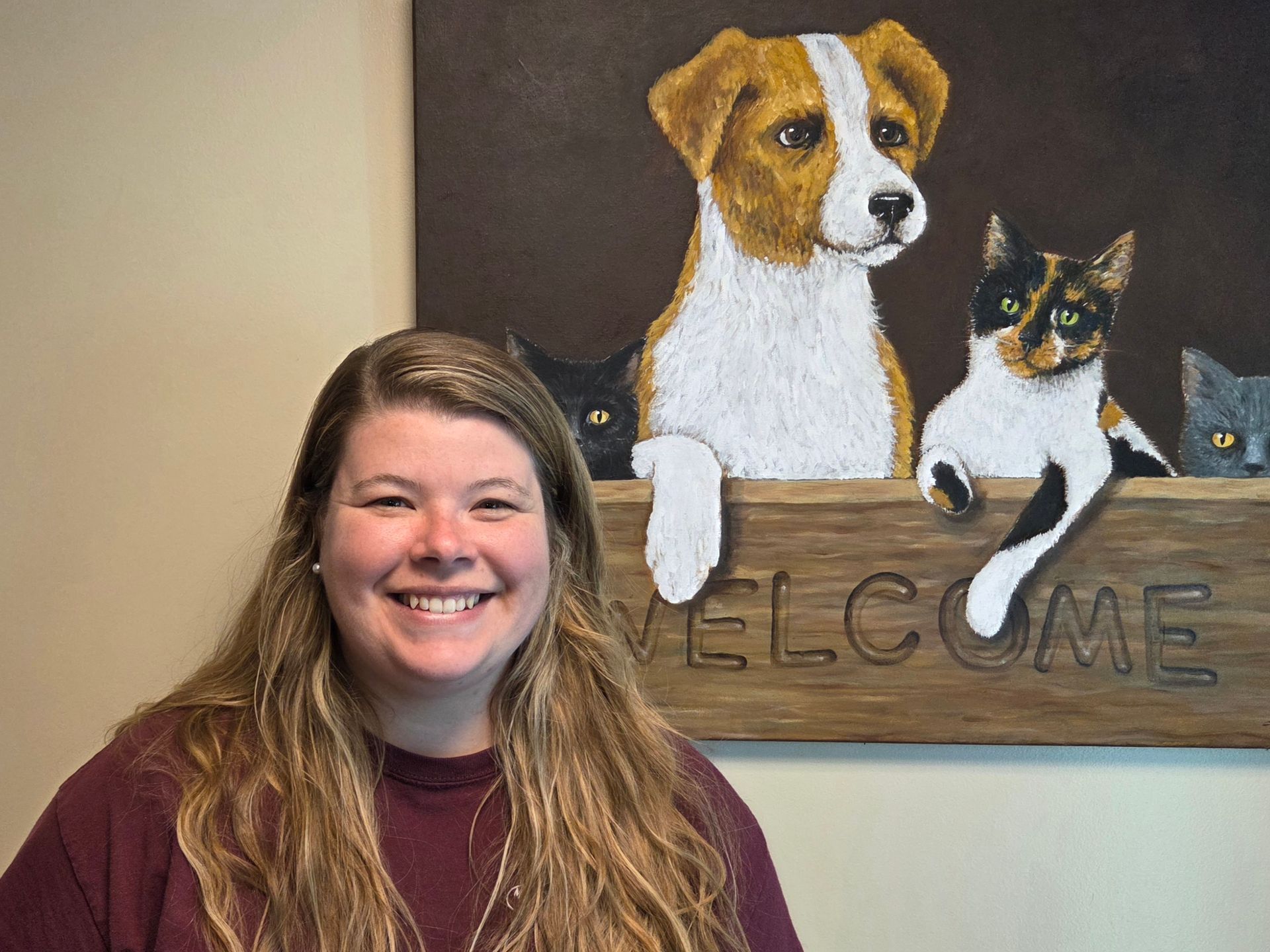 Smiling woman beside a painting of three pets above a wooden welcome sign