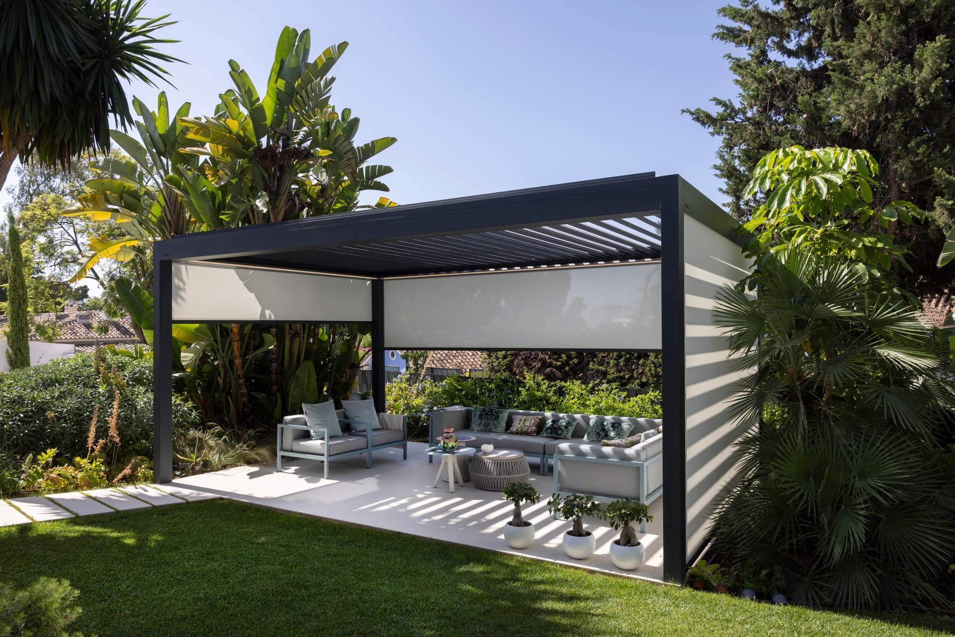 Modern outdoor patio with a black pergola, white seating, and tropical landscaping under a bright sky
