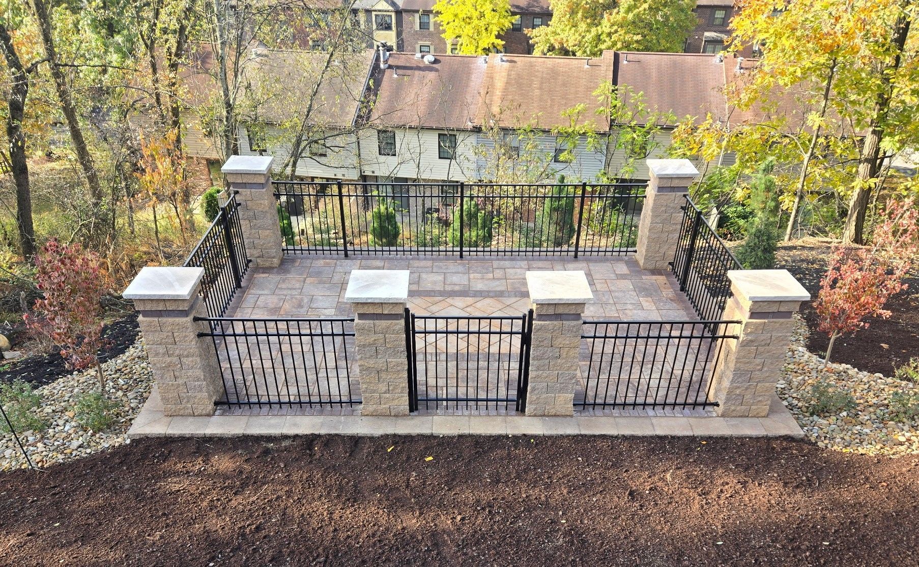Stone terrace with wrought-iron gate, brick pillars, and autumn trees overlooking a garden and house