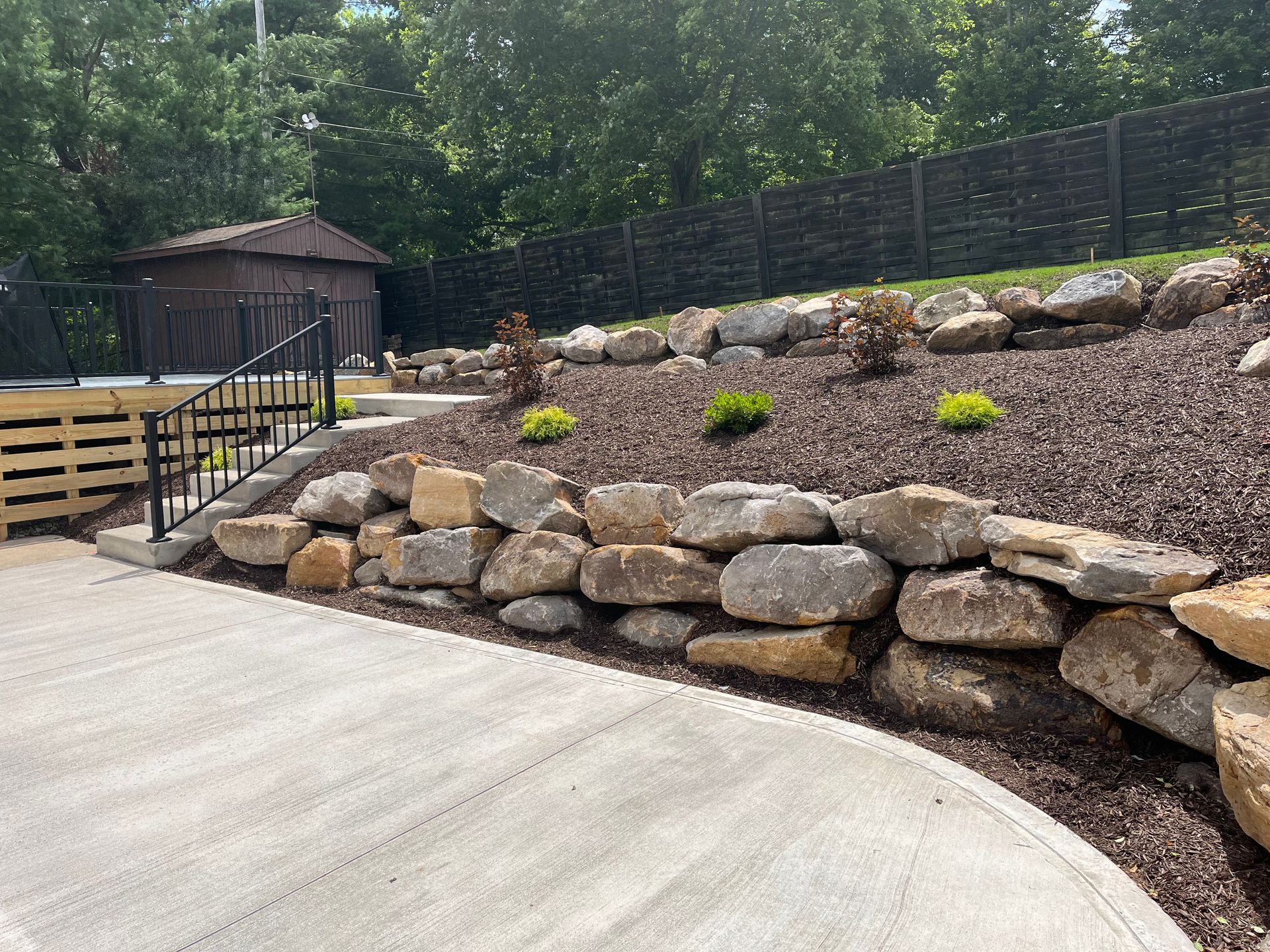 A stone wall surrounds a concrete walkway in a backyard.