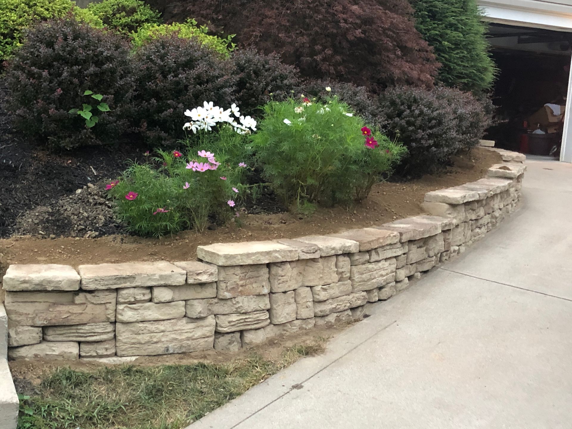 A stone wall with flowers and bushes in front of a garage.
