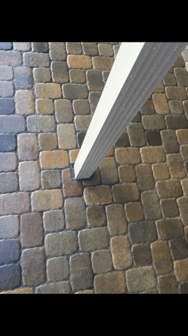 A white vertical support post resting on a patio floor made of multicolored, rectangular stone pavers.