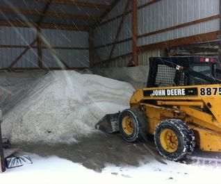 A yellow John Deere skid-steer loader parked inside a shed next to a large pile of road salt during a light snowfall.