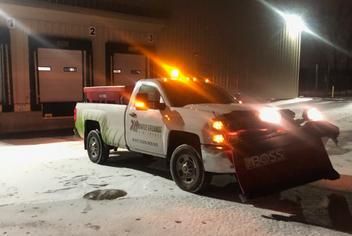 A white snowplow truck with a Boss plow and yellow caution light parked outside warehouse loading docks at night.