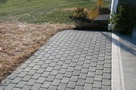 A gray paver walkway leads from a concrete driveway across a lawn with patches of straw and decorative shrubs.