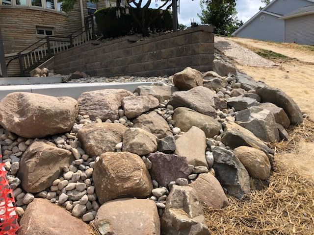 A landscaped area features large tan and grey boulders nestled among smaller river rocks next to a stone retaining wall.