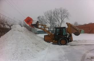 A yellow backhoe loader sits on a snowy lot, using its front bucket to pile snow into a large mound.